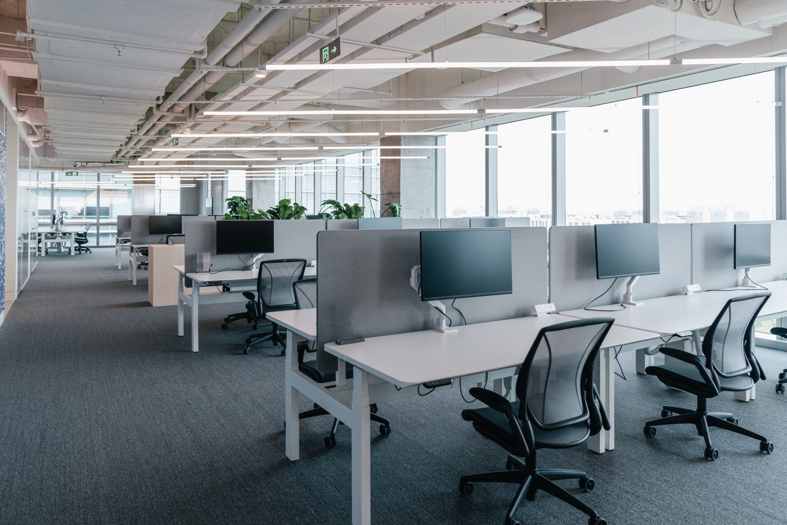 Office space featuring desks and chairs arranged in front of a large window, providing natural light.